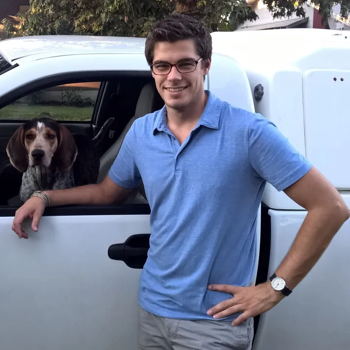 A younger Dan, smiling by a white work truck, with a hound looking out the window